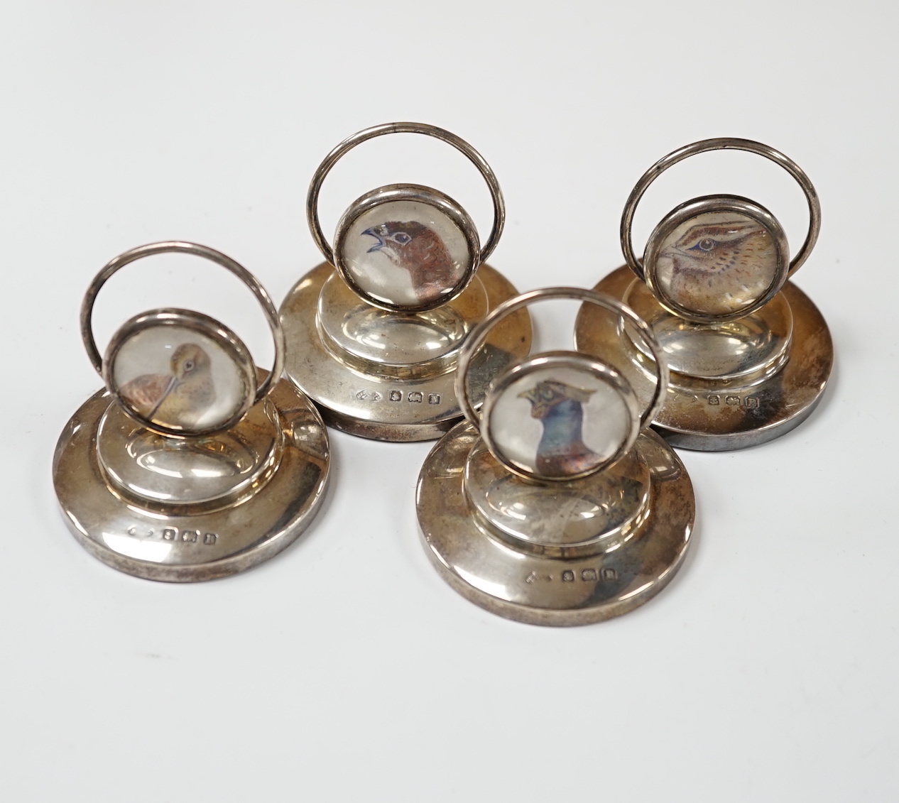 A set of four George V silver menu holders, each with inset painted panel of a wild bird, Adie Bros. Ltd, Birmingham, 1926, base diameter 5cm.